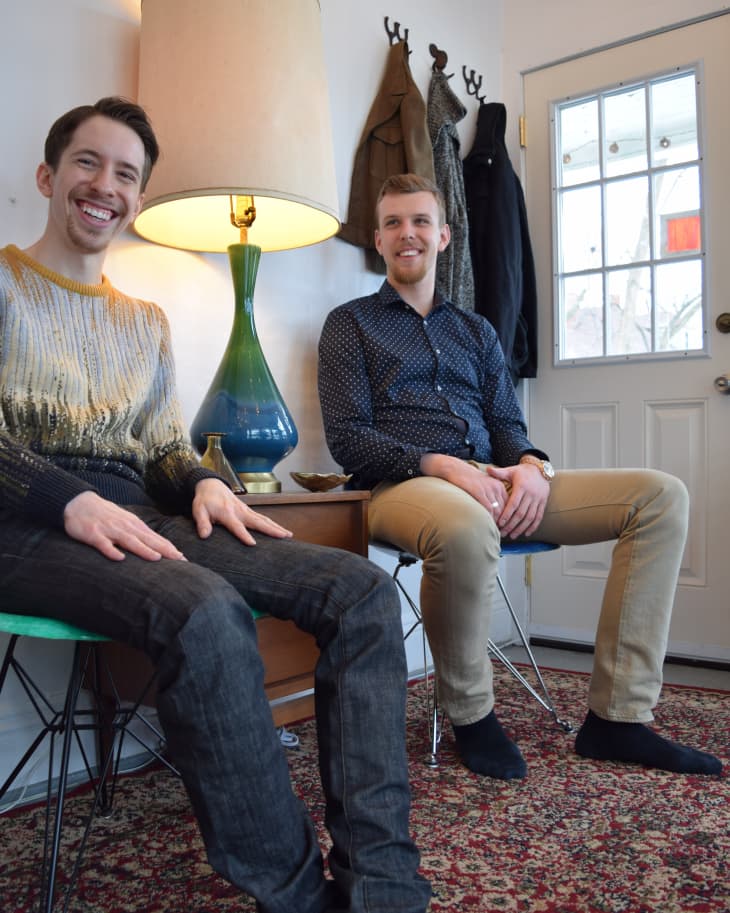 Two men sitting on chairs in a cozy room with a large lamp, patterned rug, and coats hanging by a door.