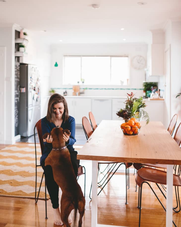 Woman playing with a brown dog in a bright kitchen, next to a wooden dining table with a fruit bowl centerpiece.