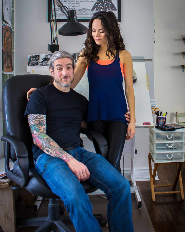 Man with tattooed arm sitting on office chair, woman standing beside him, Star Wars poster in background.
