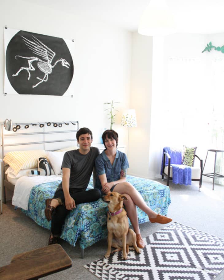 Couple sitting on a bed with a blue floral cover, dog at their feet, black and white rug, and Pegasus artwork on the wall.