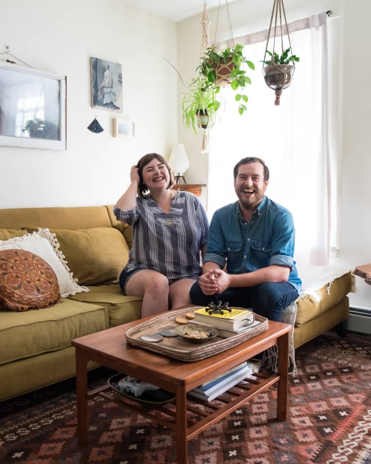 Couple sitting on a mustard sofa in a cozy living room with hanging plants, a wooden coffee table, and patterned rug.