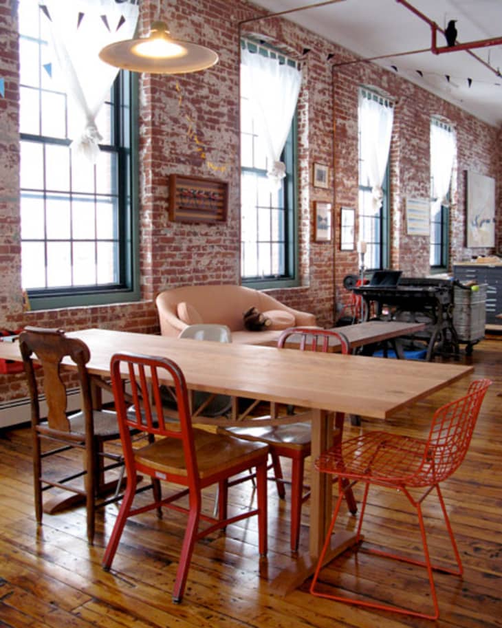 Loft-style room with exposed brick walls, wooden table, mismatched chairs, and large windows with sheer curtains.