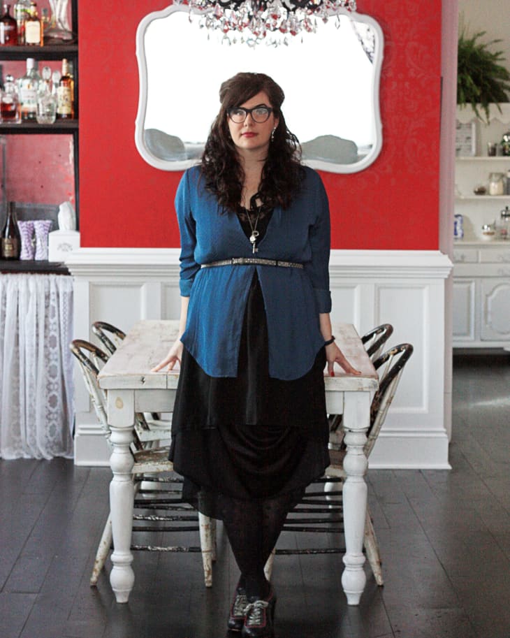 Woman in blue cardigan standing at a rustic white dining table with a black chandelier and red wall background.