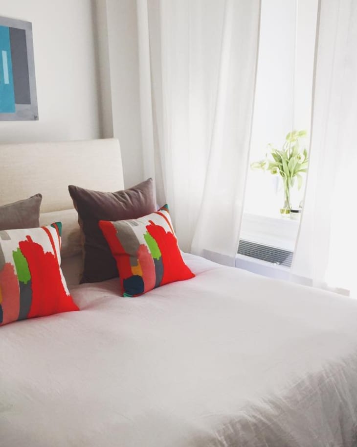Bedroom with white bedding, colorful abstract pillows, a modern painting, and a vase of flowers by the window.