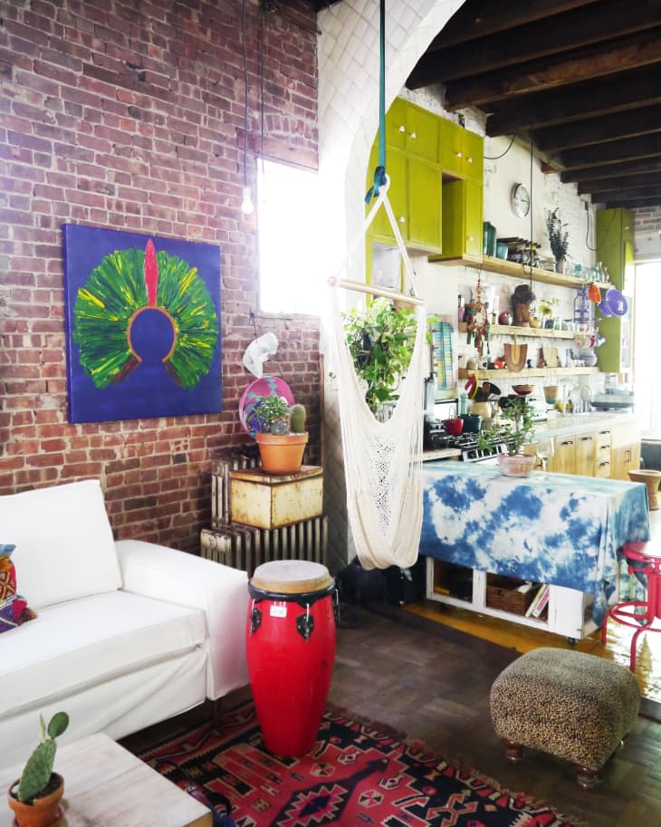 Loft-style living room with exposed brick, white sofa, red drum, hammock chair, and colorful decor.