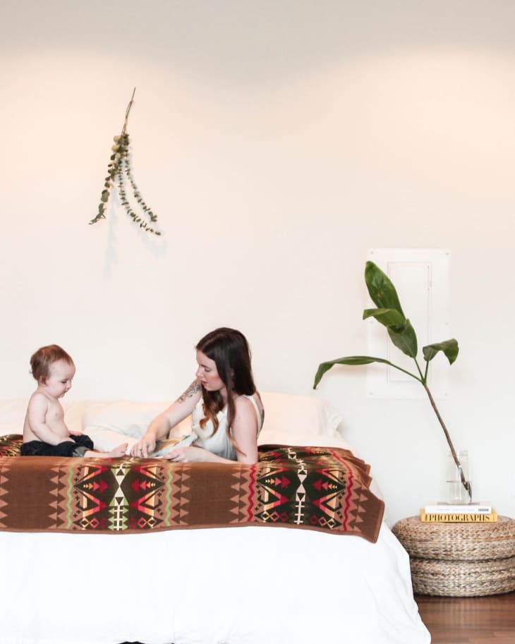 Woman and child sitting on a bed with a patterned blanket, next to a plant and books on a woven basket.
