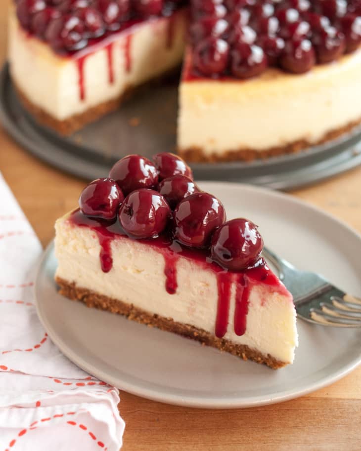 Wedge of cheesecake topped with cherries in sauce on a plate, full cherry cheesecake in background