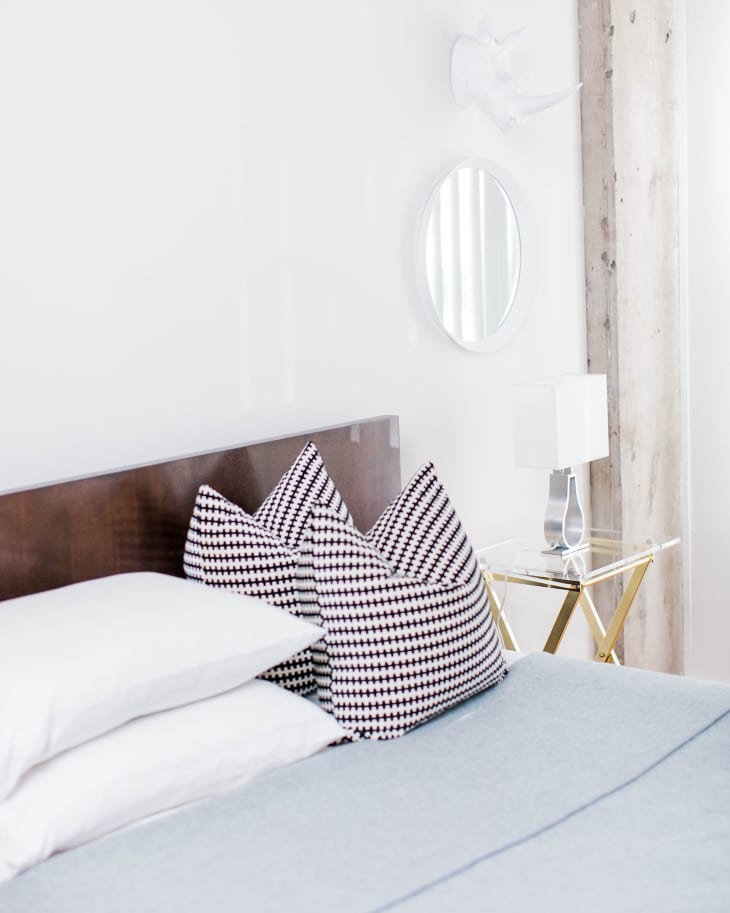 Modern bedroom with a wooden headboard, houndstooth pillows, a round mirror, and a glass side table with a lamp.