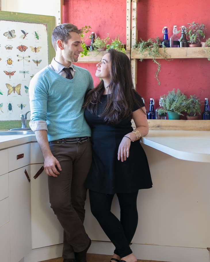 Couple smiling at each other in a kitchen with red shelves, plants, and butterfly artwork.