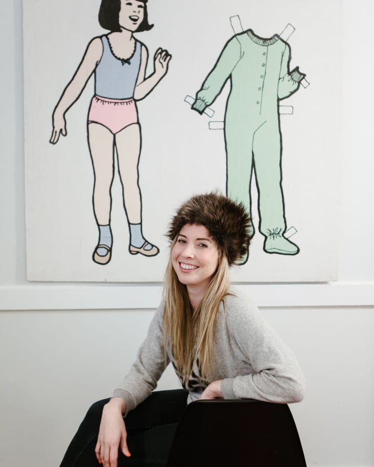 Woman in a fur hat sitting on a chair, smiling, with a paper doll art piece in the background.