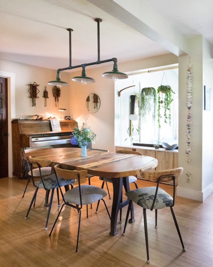 Dining room with wooden table, four chairs, hanging lights, piano, and plants by the window.
