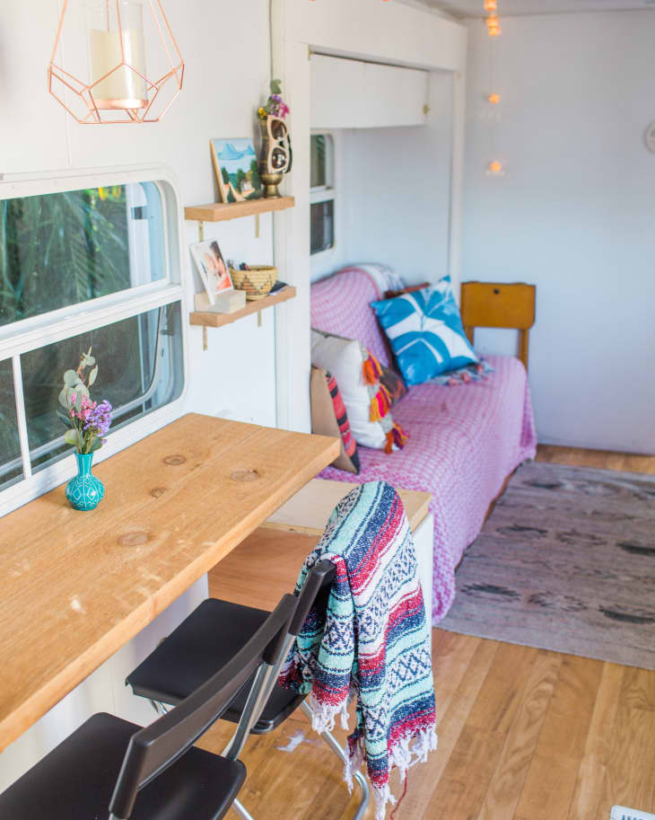 Camper interior with wooden table, black chairs, colorful throw, shelves with decor, and a pink sofa with vibrant pillows.