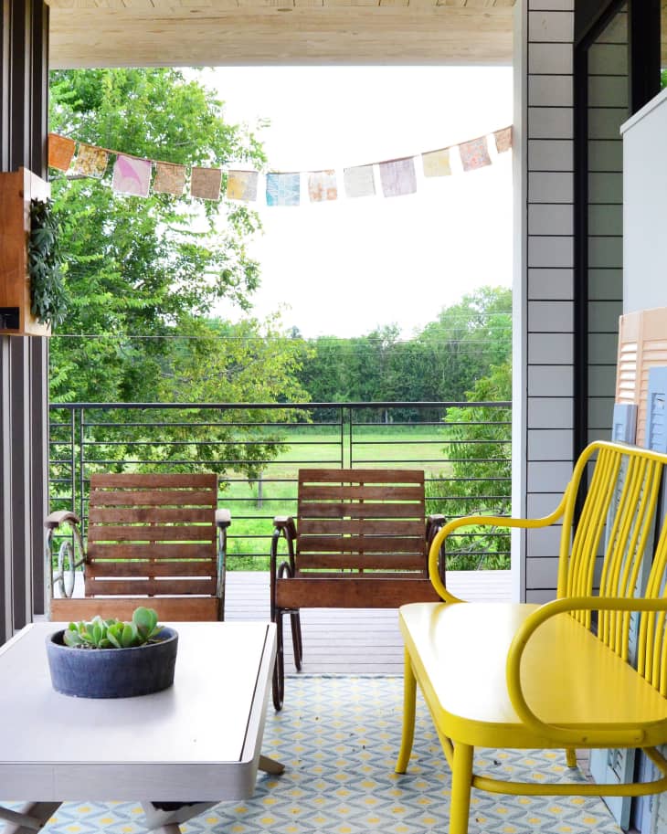 Covered patio with yellow bench, wooden chairs, potted succulents, and colorful flags overlooking a green field.