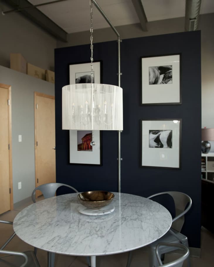 Round marble dining table with metal chairs, modern chandelier, and framed art on a dark accent wall.