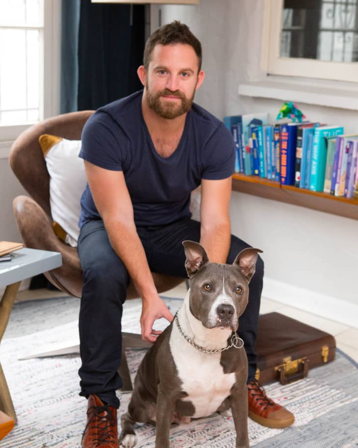 Man sitting on a chair with a gray and white dog, books on a shelf in the background.
