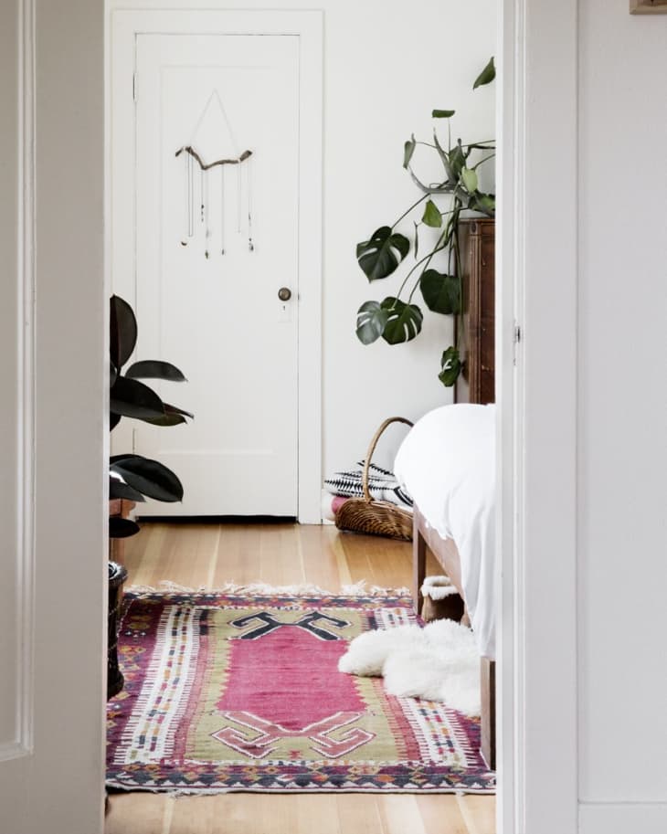 Bedroom with a colorful patterned rug, large leafy plants, and a white bedspread.