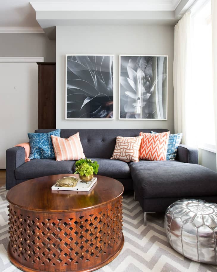 Gray sectional sofa with colorful pillows, wooden coffee table, silver pouf, and abstract black-and-white wall art.
