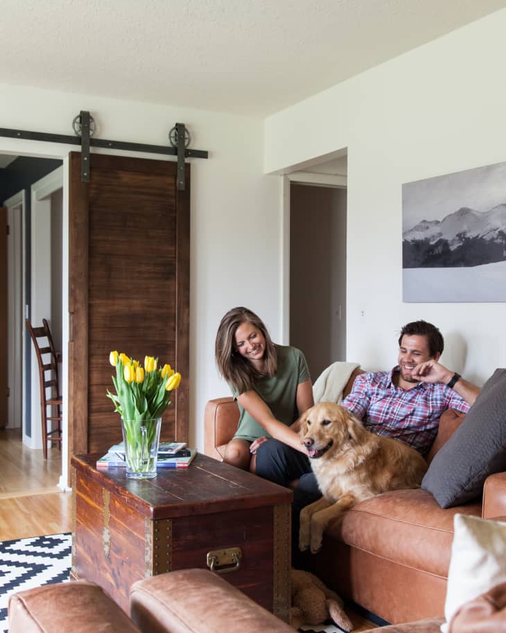 Couple sitting on a brown leather sofa with a golden retriever, next to a wooden chest and yellow tulips in a vase.