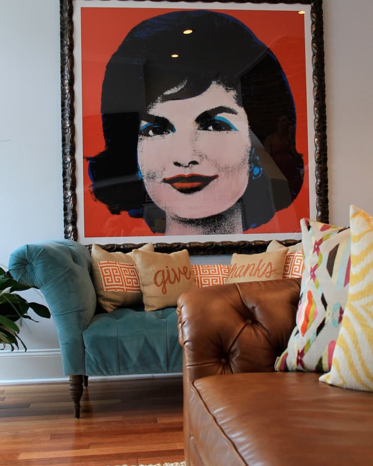 Framed pop art portrait above a teal sofa with decorative pillows and a brown leather chair in the foreground.