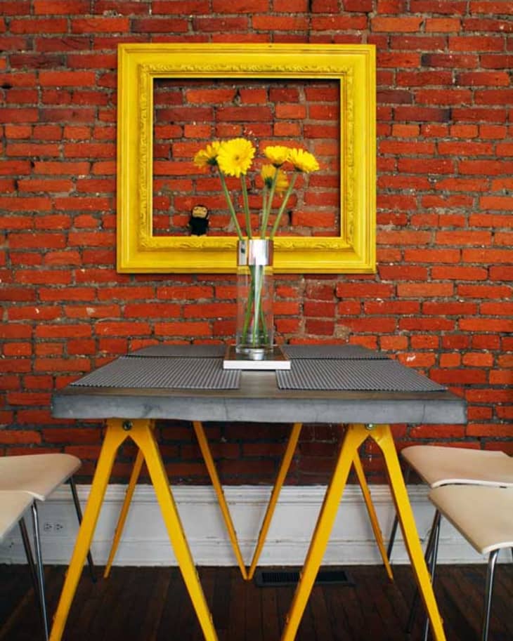 Yellow-framed mirror on red brick wall above table with yellow legs and vase of yellow flowers.