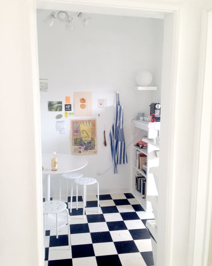 Small kitchen with black and white checkered floor, white stools, wall art, striped apron, and shelves with books and coffee maker.