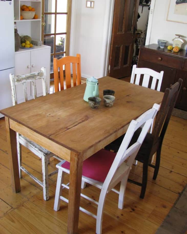 Rustic wooden dining table with mismatched chairs, a mint green teapot, and cups in a cozy kitchen setting.