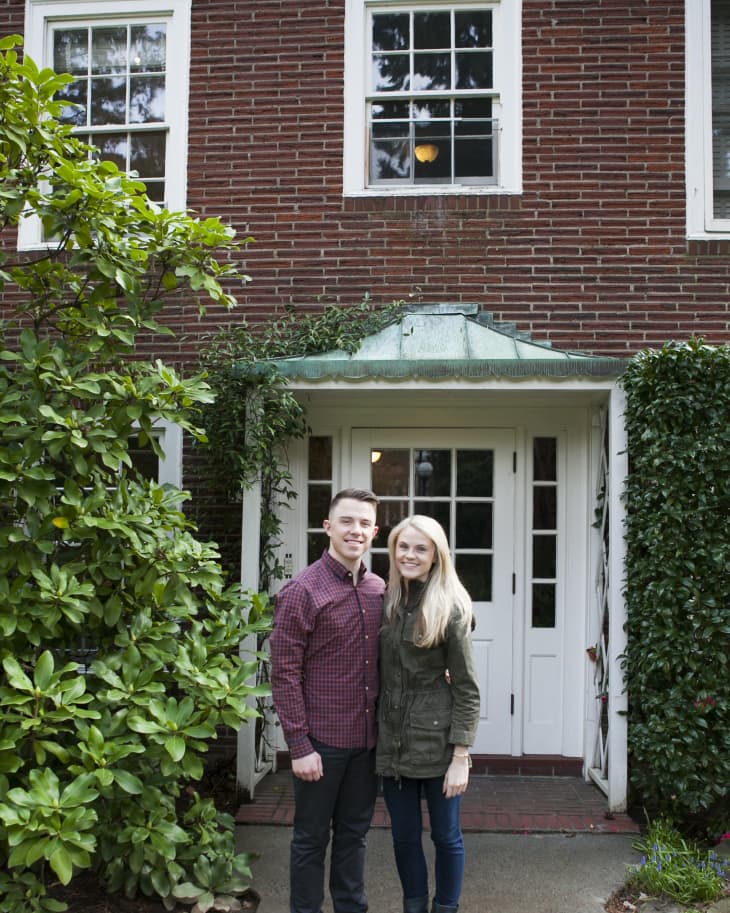 Couple standing in front of a brick house with white-framed windows and a green canopy, surrounded by greenery.