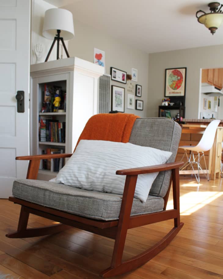 Mid-century modern rocking chair with gray cushions, orange throw, and white pillow in a cozy living room with wooden floor.