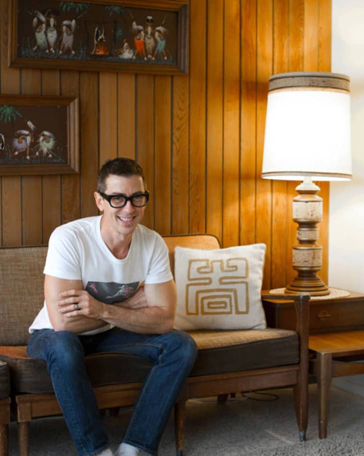 Man in glasses sitting on a vintage sofa with a geometric pillow, next to a wooden lamp and paneled wall with framed art.