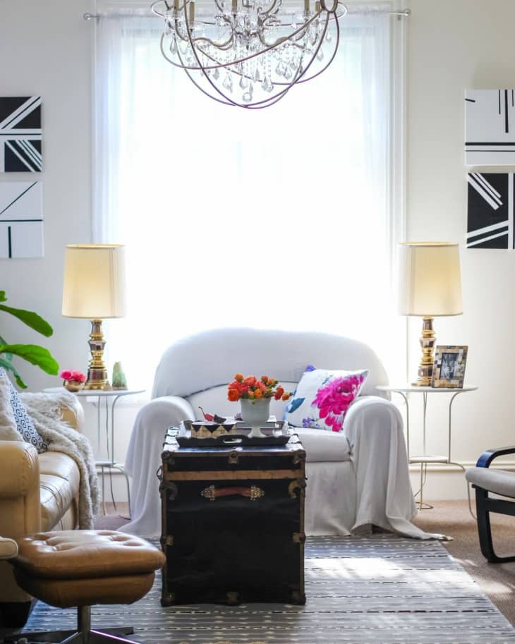 Living room with a white armchair, floral pillow, vintage trunk coffee table, and chandelier.