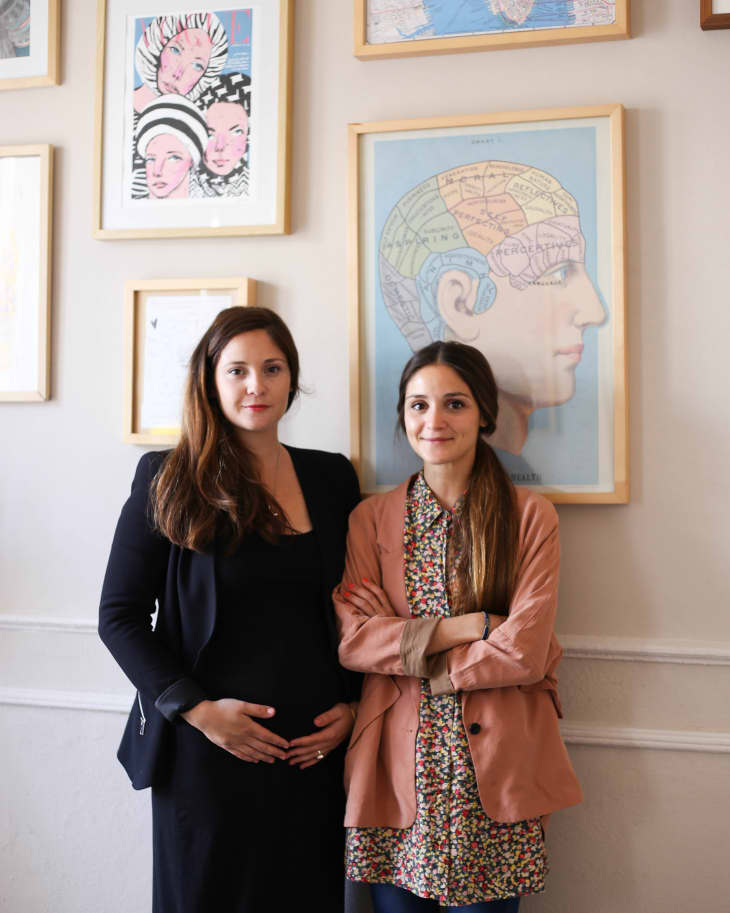 Two women standing in front of a wall with framed art, including a brain diagram and abstract portraits.