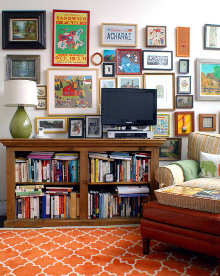 Living room with a gallery wall, wooden bookshelf, TV, green lamp, striped armchair, and orange patterned rug.