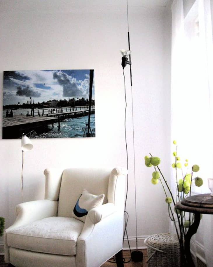 Cozy reading nook with a white armchair, floor lamp, and wall art of a pier. Green floral decor adds a fresh touch.