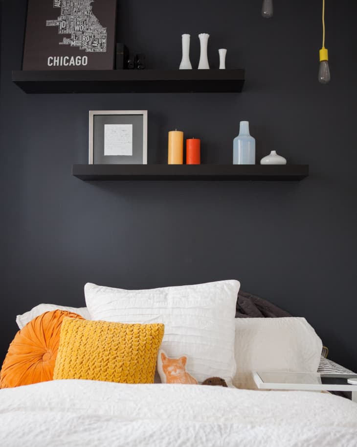 Dark bedroom wall with floating shelves, Chicago map art, vases, colorful candles, and a bed with white and yellow pillows.