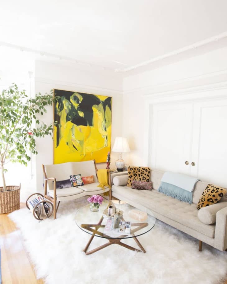 Living room with beige sofa, glass coffee table, abstract yellow painting, and a large potted plant.