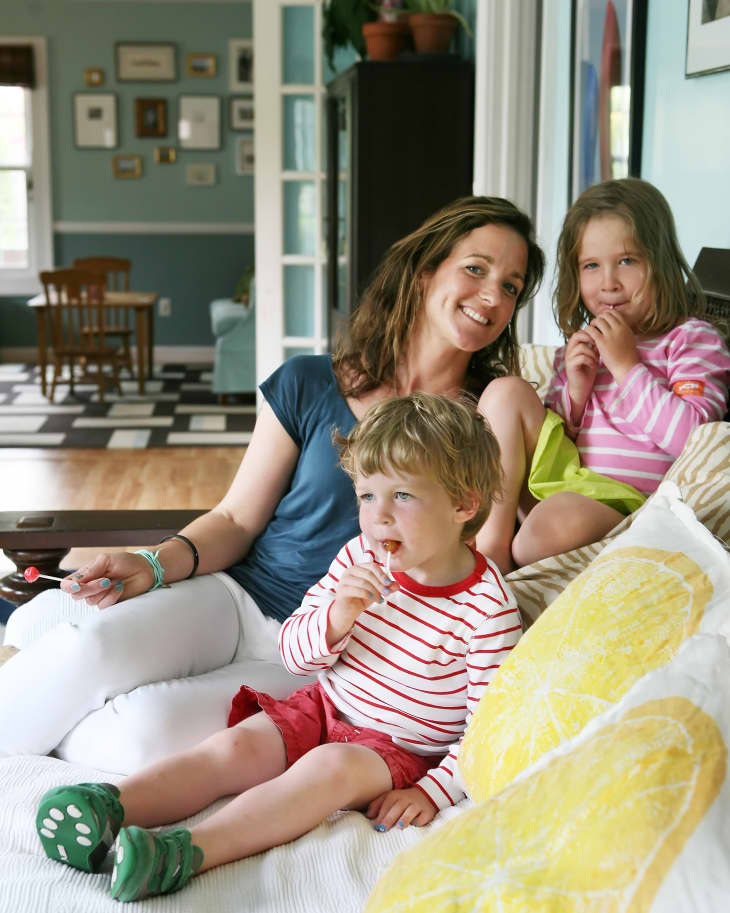 Woman and two children sitting on a bed with lollipops, in a colorful room with framed art and plants.