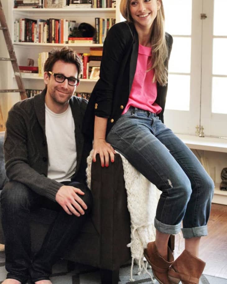 Man and woman in casual attire sitting in a cozy living room with bookshelves and a patterned rug.