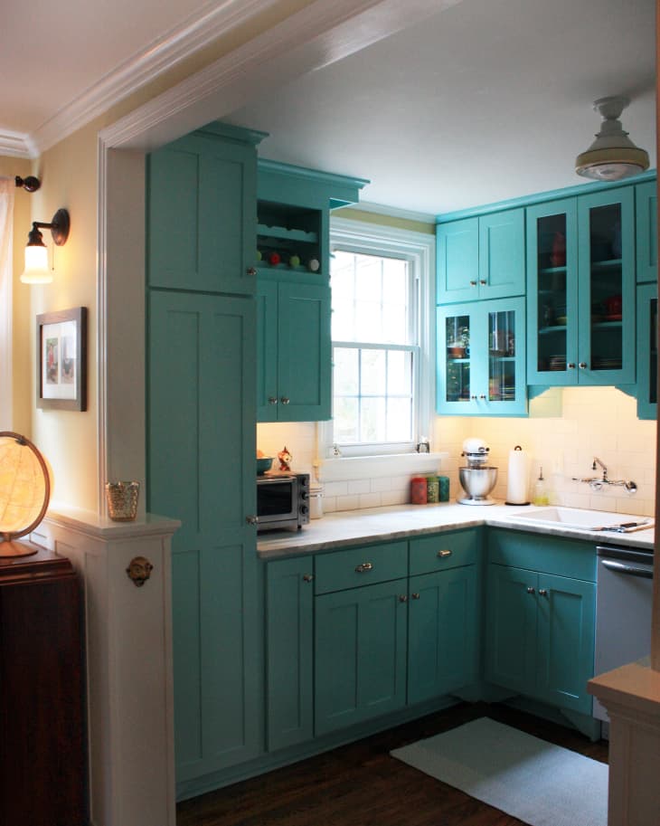 Small kitchen with turquoise cabinets, white countertops, a window, and wooden floor. Mixer and toaster on counter.