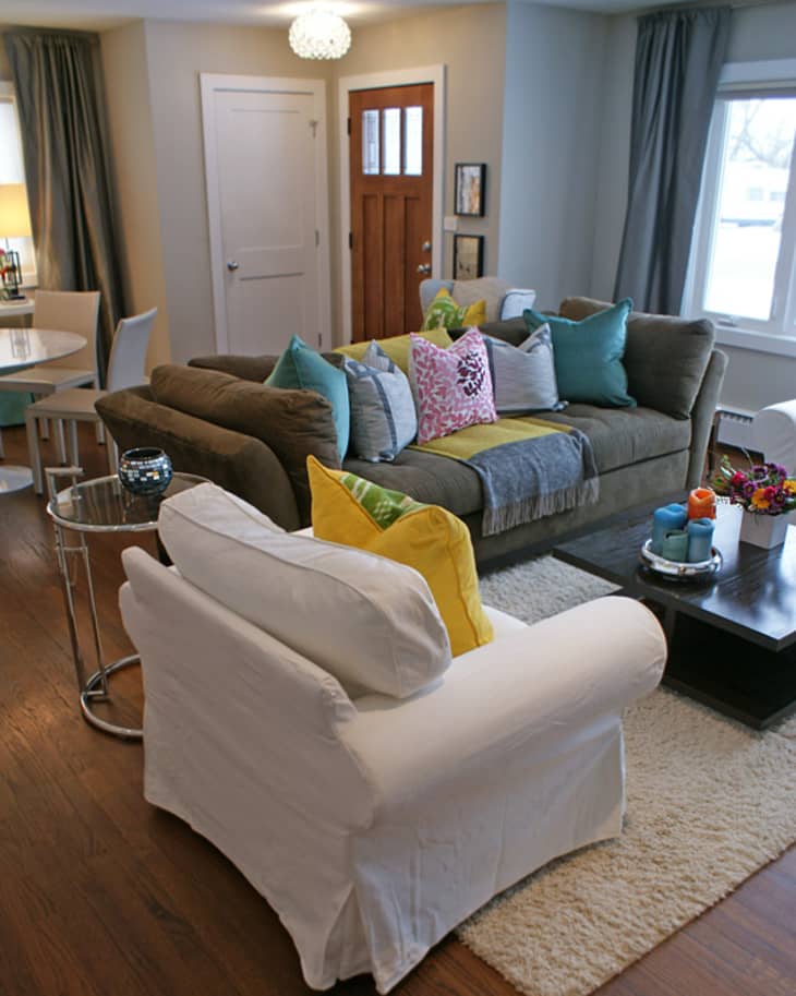 Living room with a brown sofa, colorful pillows, white armchair, and a dark coffee table with candles and flowers.