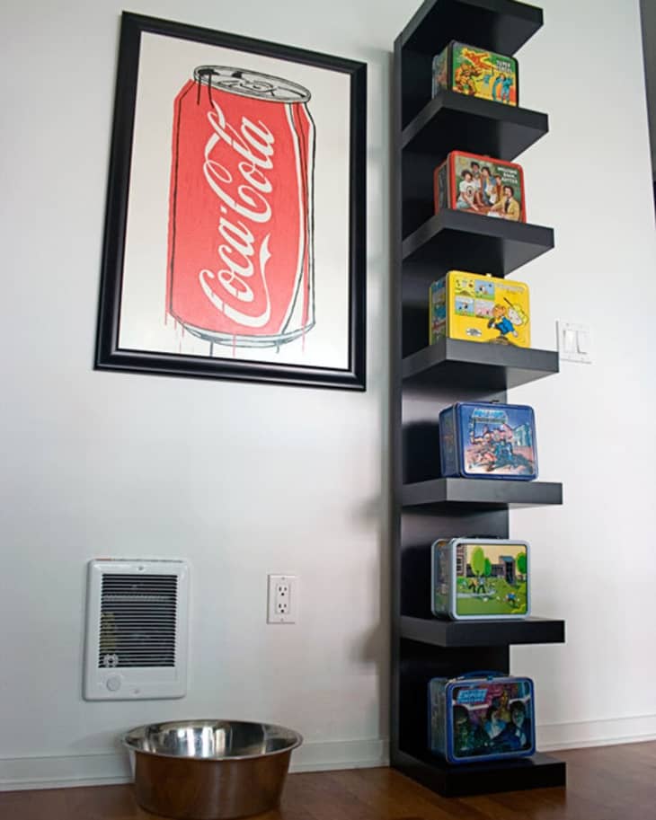 Framed Coca-Cola can art above a vertical shelf with vintage lunchboxes, next to a metal pet bowl on wooden floor.