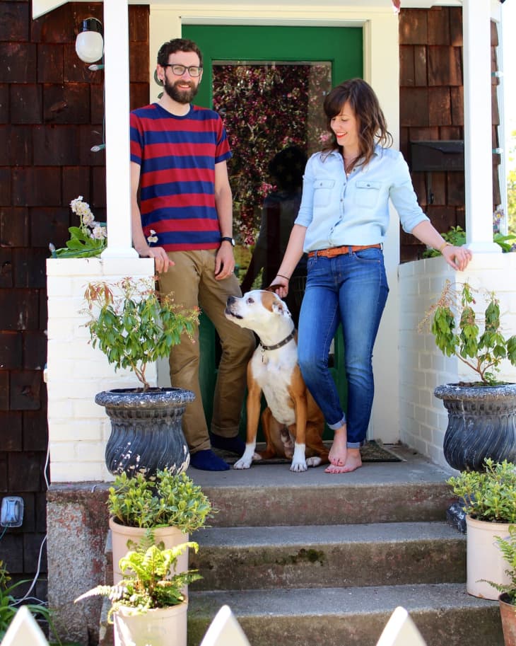 Couple with a dog on porch steps, surrounded by potted plants and a green door.