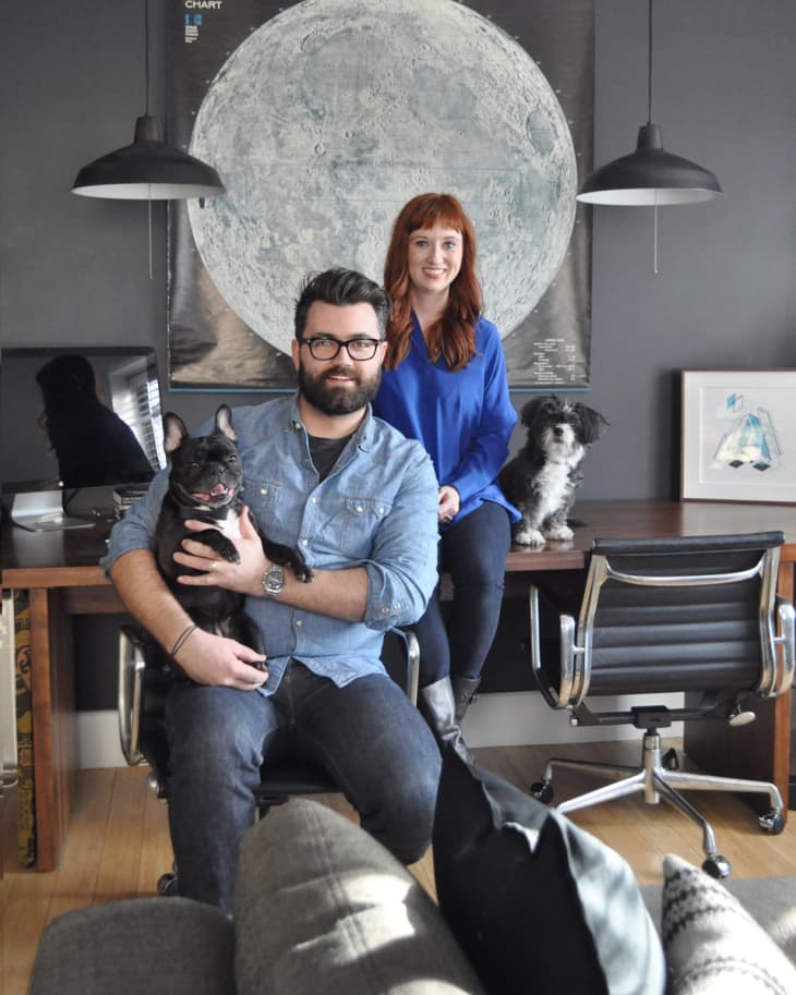 Couple with two dogs in a modern office, sitting by a desk with a large moon poster and black pendant lights.