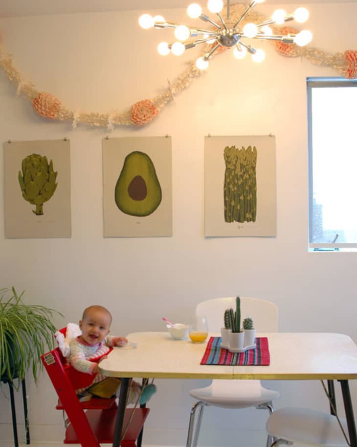Baby in a red high chair at a dining table with cactus decor, artichoke, avocado, and asparagus wall art.