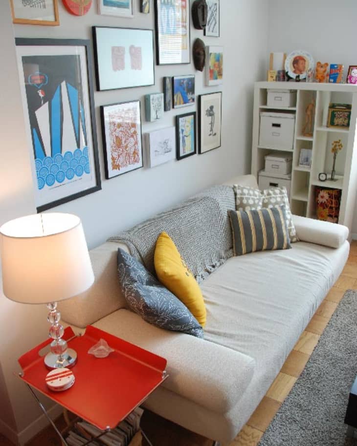 Cozy living room with a white sofa, colorful cushions, gallery wall art, and a red side table with a lamp.