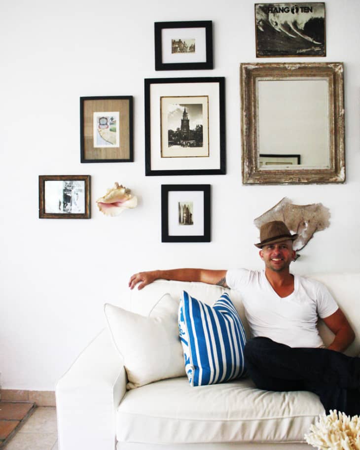 Man in a hat sitting on a white sofa with striped pillows, surrounded by framed art and a mirror on the wall.