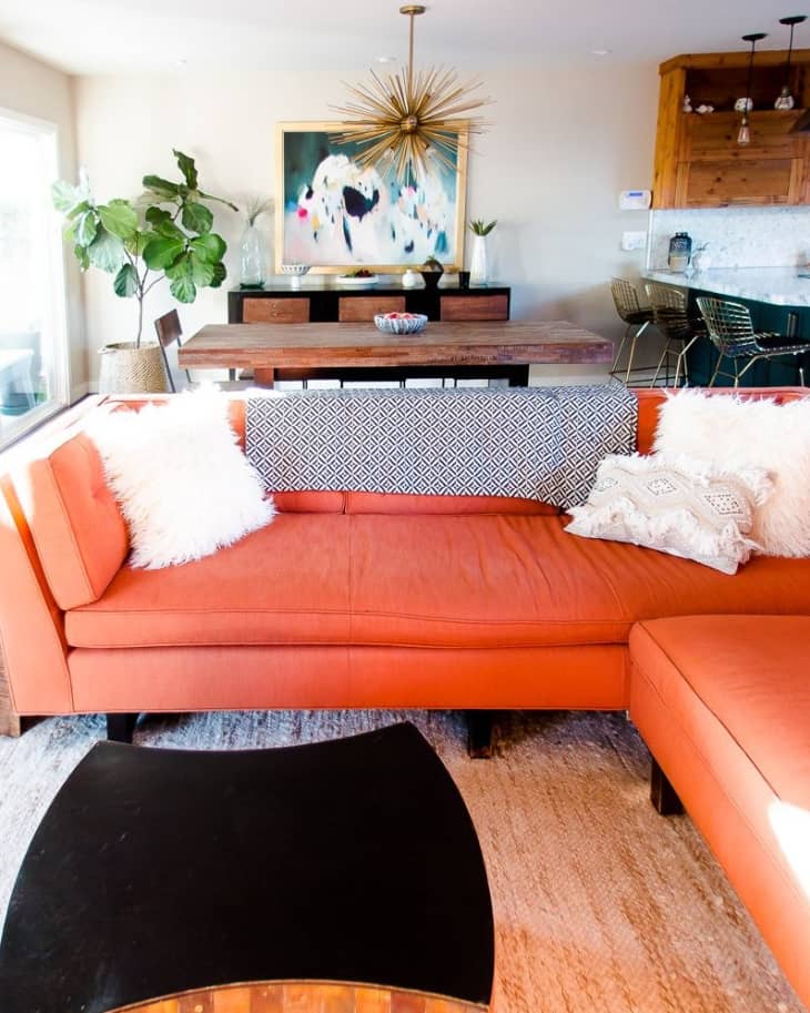Orange sectional sofa with patterned throw, fluffy pillows, wooden coffee table, and dining area with abstract art and chandelier.