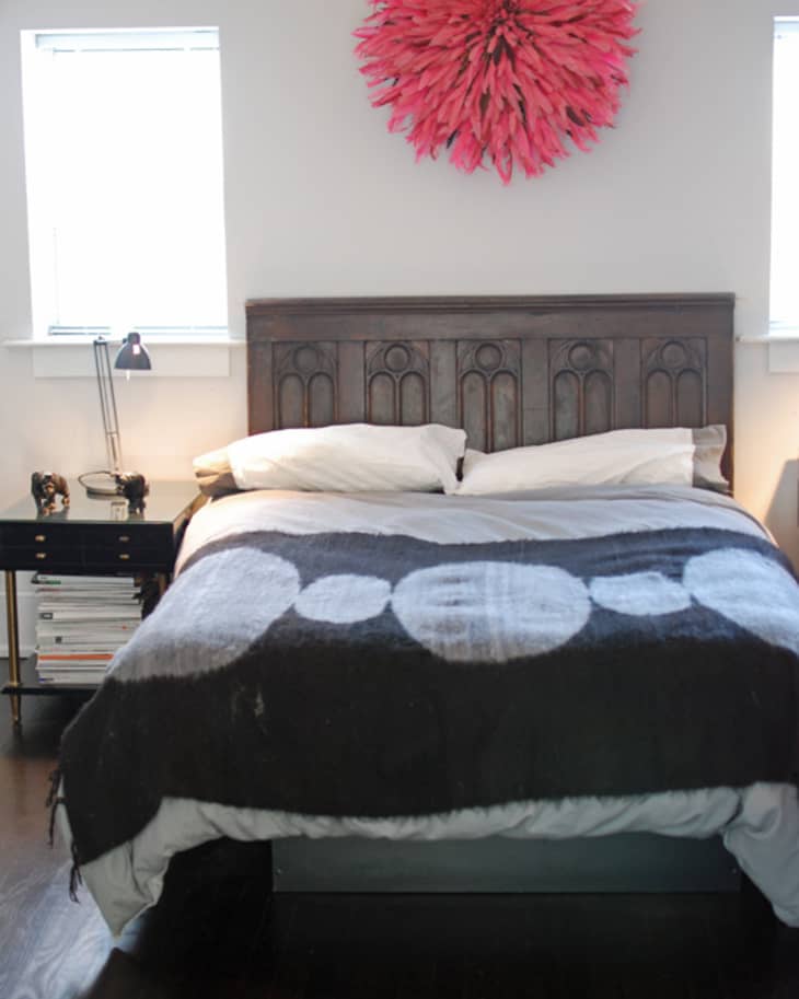Bedroom with dark wood headboard, black and white patterned blanket, red feather wall decor, and bedside table with lamp.