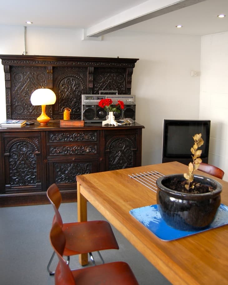 Dining area with wooden table, brown chairs, ornate dark cabinet, table lamp, boombox, and potted plant.