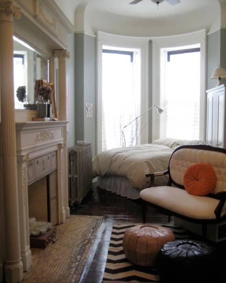 Cozy bedroom with a fireplace, bay windows, a bed, and an armchair with orange and black cushions on a chevron rug.