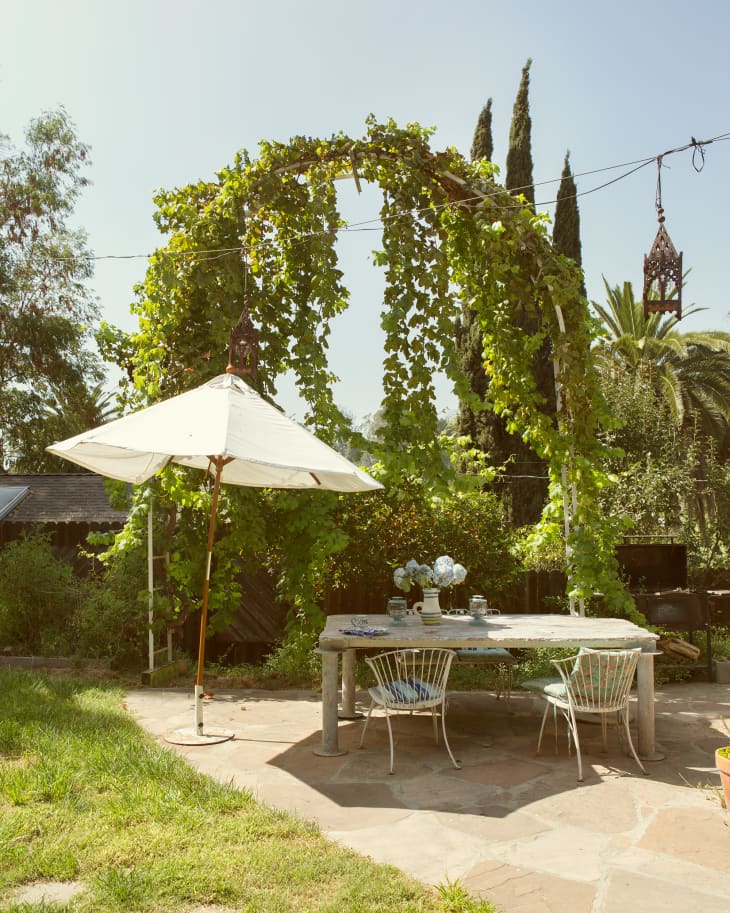 Outdoor patio with a wooden table, white chairs, large umbrella, and vine-covered archway.
